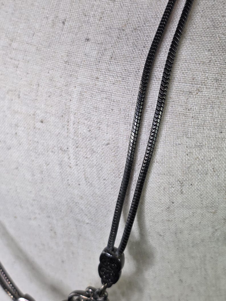 May include: Close-up of a black metal necklace with a snake chain design. The necklace has a detailed pendant and a textured clasp. The background is a light-coloured fabric.