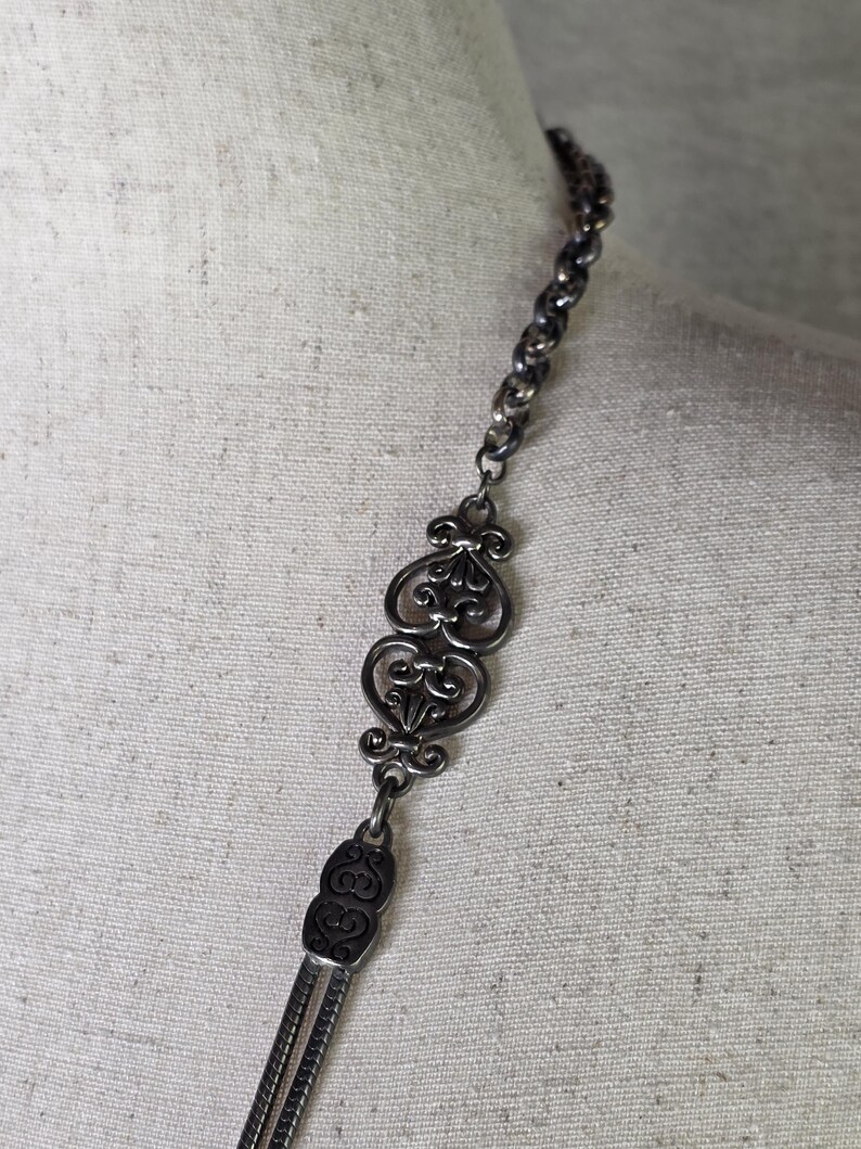 May include: A silver necklace with an elaborate, filigree design. The necklace has a chain, decorative centrepieces, and a tassel-like drop. The intricate details and vintage style create a classic look.
