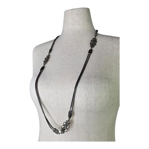 May include: A long, gunmetal necklace displayed on a mannequin. The necklace has decorative, ornate links and a snake chain design. The necklace is a dark, metallic grey and hangs down the mannequin's torso.