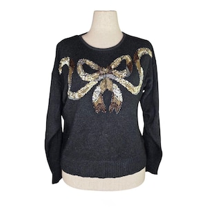 May include: A dark gray sweater with a sequined bow design. The bow features gold, brown, and white sequins. The sweater has long sleeves and a crew neck. The garment is displayed on a mannequin.