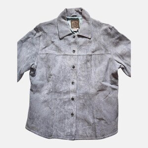 May include: A gray suede button-up shirt with a collared neckline. The shirt has a worn, textured appearance and features a series of dark buttons down the front. The shirt has a label on the inside collar.