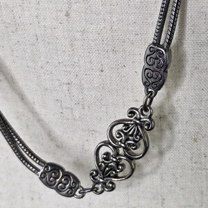 May include: A silver necklace with an intricate design. The necklace has a double strand chain and decorative connectors with swirling patterns and a fleur-de-lis motif. The metal has a slightly aged appearance.