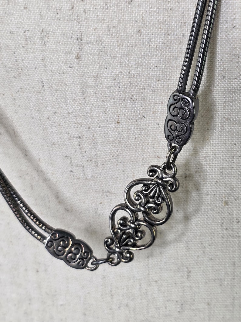 May include: A silver necklace with an intricate design. The necklace has a double strand chain and decorative connectors with swirling patterns and a fleur-de-lis motif. The metal has a slightly aged appearance.