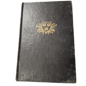 Puede incluir: Un libro de tapa dura negro con un dise&ntilde;o en relieve dorado en la portada. El dise&ntilde;o presenta un escudo con tres coronas y un par de espadas cruzadas.