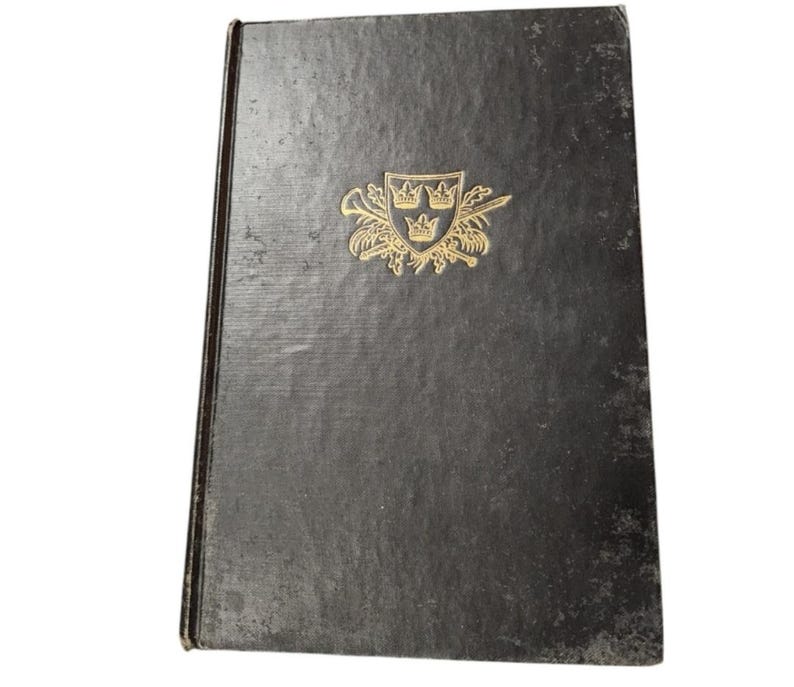 Puede incluir: Un libro de tapa dura negro con un dise&ntilde;o en relieve dorado en la portada. El dise&ntilde;o presenta un escudo con tres coronas y un par de espadas cruzadas.