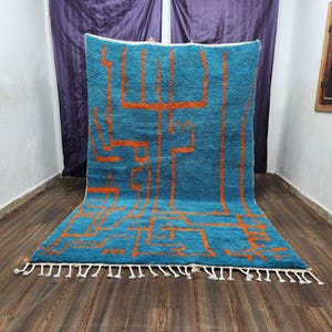 May include: A turquoise rug with an abstract orange geometric design. The rug has a thick pile and a fringe of white tassels along the bottom edge. The rug is displayed on a wooden floor.