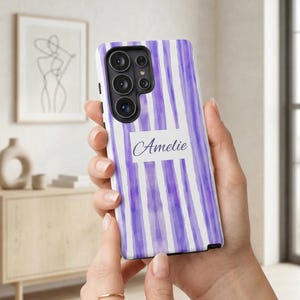 Puede incluir: Funda de teléfono con diseño de acuarela de rayas verticales moradas y blancas. El nombre "Amelie" está escrito en cursiva sobre un rectángulo blanco. La funda tiene un módulo de cámara con múltiples lentes.