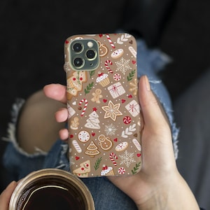 Puede incluir: Una funda de teléfono con un patrón navideño festivo. La funda presenta un fondo marrón con ilustraciones de hombres de jengibre, bastones de caramelo, copos de nieve y regalos envueltos en rojo, blanco y marrón. La funda está en una mano.