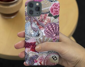 May include: A decorative phone case with a collage of beaded embellishments in pink, purple, and cream colors. The design includes a rose, a butterfly, and an eight ball.