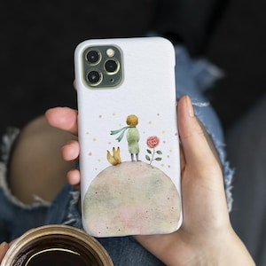 May include: White phone case featuring a watercolor illustration of The Little Prince. The design includes the prince, a fox, a rose, and a small planet. The case has a glossy finish and is designed for a smartphone.