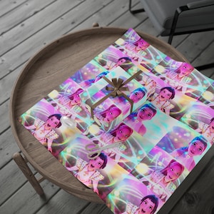 Puede incluir: Una caja de regalo envuelta con una cinta marrón se encuentra sobre una hoja de papel de regalo estampado. El papel y la caja presentan un diseño repetido de un personaje de dibujos animados con un atuendo rosa y blanco, sobre un fondo de líneas coloridas y brillantes.