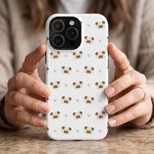 Puede incluir: Funda de teléfono blanca con un patrón repetido de caras de dibujos animados de pug y huellas de patas. Los pugs tienen caras color canela, ojos y narices negros. Las huellas de las patas son de color marrón claro.