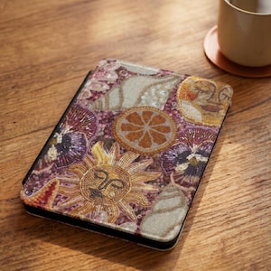 Puede incluir: Funda decorativa para tableta con un diseño bordado con un sol, conchas marinas y elementos florales en tonos dorados, morados y blancos. La funda está sobre una superficie de madera, con una taza y un posavasos en el fondo.