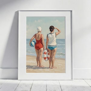 Op de afbeelding: Een ingelijste prent van twee vrouwen op het strand. Eén draagt een rood badpak en houdt een blauw-witte bal vast. De ander draagt een wit en blauw badpak en houdt een rood-witte reddingsboei vast. Ze staan op het zand en kijken naar de oceaan.