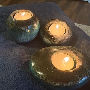May include: Three ceramic tealight candle holders with lit candles. The holders are in shades of green and brown, with a speckled glaze. The candles emit a warm, inviting glow, creating a relaxing ambiance.