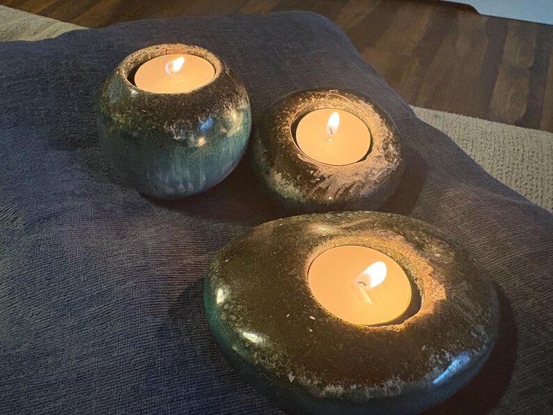 May include: Three ceramic tealight candle holders with lit candles. The holders are in shades of green and brown, with a speckled glaze. The candles emit a warm, inviting glow, creating a relaxing ambiance.
