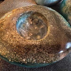 May include: A unique, handcrafted teal and brown resin candle holder. The circular design features a central opening and is embedded with small stones, creating a textured, decorative piece. The resin has a glossy finish.
