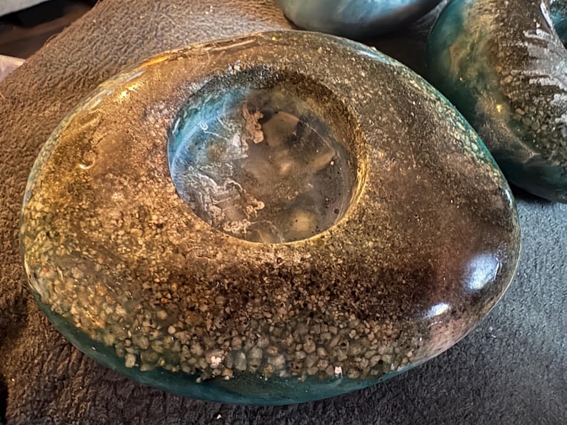 May include: A unique, handcrafted teal and brown resin candle holder. The circular design features a central opening and is embedded with small stones, creating a textured, decorative piece. The resin has a glossy finish.