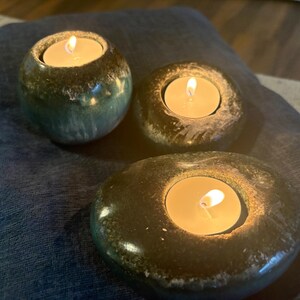 May include: Three tealight candle holders with lit candles. The holders are made of a dark, speckled material, with a rounded shape. The candles emit a warm, inviting glow, creating a cozy atmosphere. The holders are placed on a blue surface.