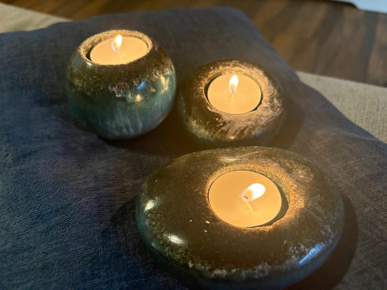 May include: Three tealight candle holders with lit candles. The holders are made of a dark, speckled material, with a rounded shape. The candles emit a warm, inviting glow, creating a cozy atmosphere. The holders are placed on a blue surface.