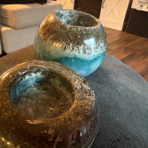 May include: Two decorative, glazed ceramic candle holders. One is a rounded shape with a teal and brown color scheme. The other is a ring shape with a similar color palette. Both have a glossy finish and are placed on a gray surface.