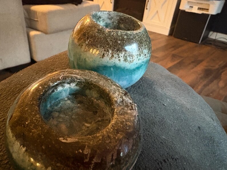 May include: Two decorative, glazed ceramic candle holders. One is a rounded shape with a teal and brown color scheme. The other is a ring shape with a similar color palette. Both have a glossy finish and are placed on a gray surface.