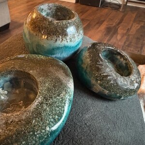 May include: Three decorative candle holders with a speckled, textured finish. The holders are in shades of brown, teal, and blue. One is round, one is oval, and the other is a sphere. The candle holders are on a gray surface.