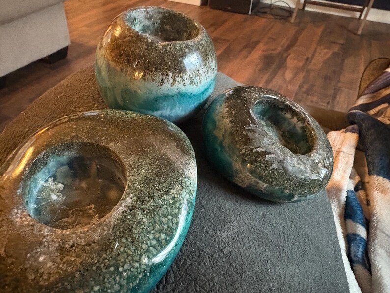 May include: Three decorative candle holders with a speckled, textured finish. The holders are in shades of brown, teal, and blue. One is round, one is oval, and the other is a sphere. The candle holders are on a gray surface.