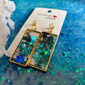 May include: Gold-framed rectangular earrings with a clear resin interior filled with teal glitter, blue flowers, and black silhouettes. The earrings hang from gold hooks. A white card with the text "Handmade with love" is behind the earrings.