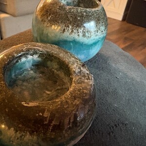 May include: Two handcrafted ceramic candle holders. One is a low, open bowl shape, the other is a round vase shape. Both have a mottled brown and turquoise glaze, creating a unique, textured appearance. The candle holders are suitable for home decor.