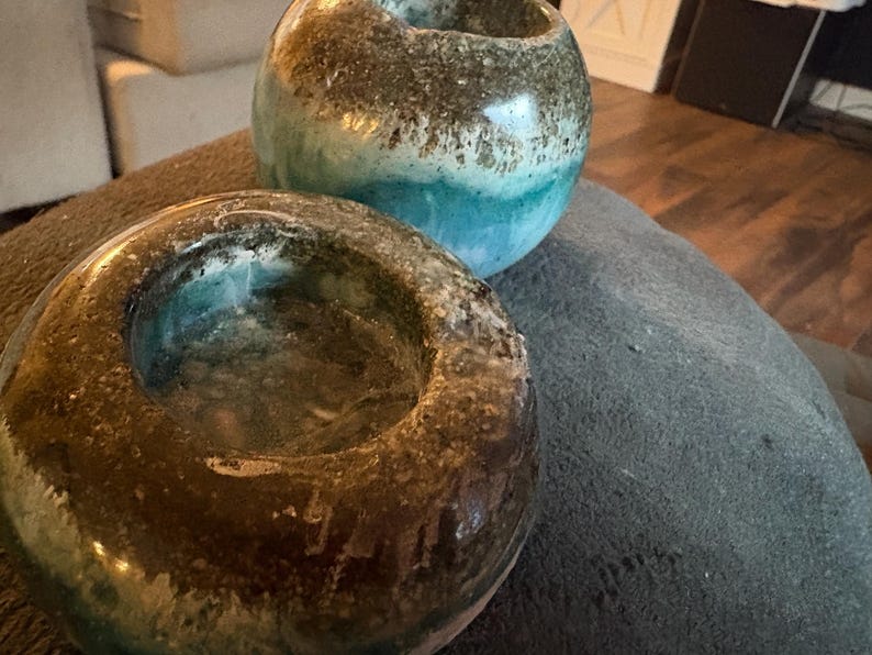 May include: Two handcrafted ceramic candle holders. One is a low, open bowl shape, the other is a round vase shape. Both have a mottled brown and turquoise glaze, creating a unique, textured appearance. The candle holders are suitable for home decor.
