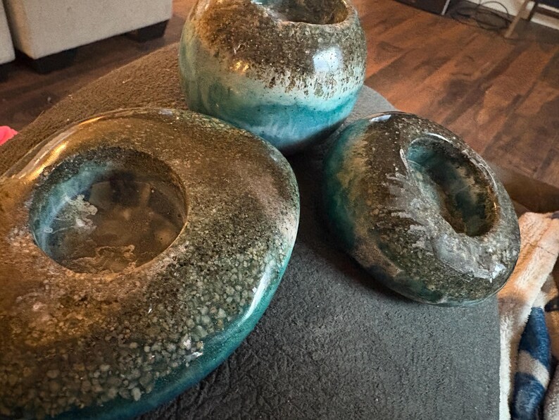 May include: Three decorative resin pieces in shades of teal, brown, and clear. The pieces include two oval-shaped candle holders and a small round vase. The resin has a textured, speckled appearance, creating a unique aesthetic.