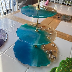 May include: A three-tiered serving tray with a gold metal stand. Each tier features a unique resin design with blue, teal, and tan colours, resembling an ocean scene with embedded seashells and starfish. The edges of each tier have a wavy, organic shape.