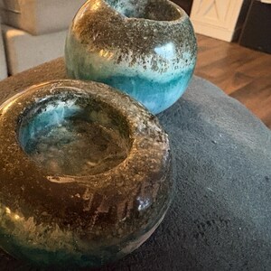 May include: Two handcrafted ceramic candle holders. The candle holders are round with a dark brown and teal glaze. The candle holders are sitting on a gray surface. The candle holders are decorative and can be used for home decor.