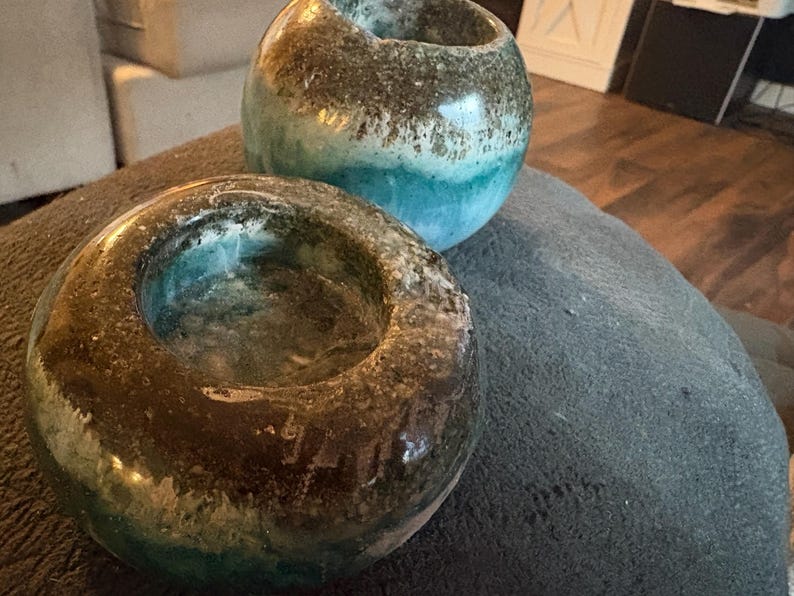 May include: Two handcrafted ceramic candle holders. The candle holders are round with a dark brown and teal glaze. The candle holders are sitting on a gray surface. The candle holders are decorative and can be used for home decor.