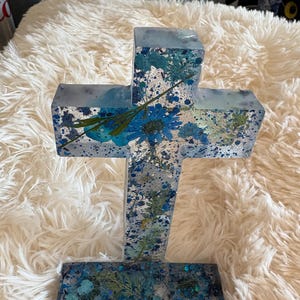 Peut inclure: Une croix décorative en résine transparente, avec des fleurs bleues et de la verdure incrustées. La croix a un motif argenté moucheté et est posée sur un fond blanc duveteux. La croix mesure environ 20 cm de haut.