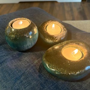 May include: Three tealight candle holders with lit candles. The holders are made of a speckled, glazed ceramic with a mix of green, brown, and blue hues. The holders are placed on a blue fabric surface.