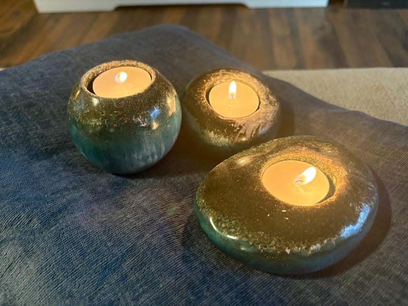 May include: Three tealight candle holders with lit candles. The holders are made of a speckled, glazed ceramic with a mix of green, brown, and blue hues. The holders are placed on a blue fabric surface.