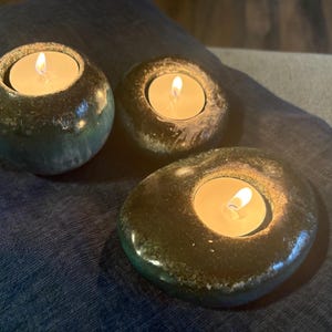 May include: Three tealight candle holders with lit candles. The holders are made of a dark, speckled material, with a glossy finish. The candle holders are different shapes and sizes, creating a unique display.