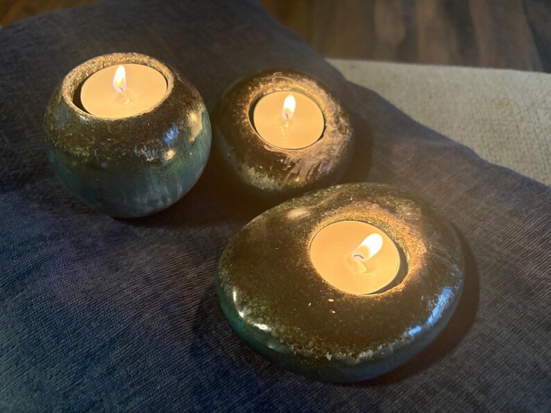 May include: Three tealight candle holders with lit candles. The holders are made of a dark, speckled material, with a glossy finish. The candle holders are different shapes and sizes, creating a unique display.