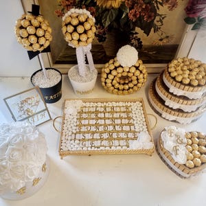 May include: A table display with a variety of gold and white themed party decorations. There are two chocolate trees, a chocolate letter board, a chocolate cake, and a chocolate heart with the letters "F & F" on it.
