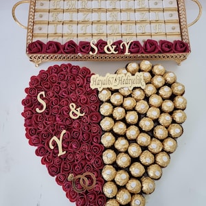 May include: A decorative arrangement featuring a heart-shaped design. One half is covered in red roses, the other with gold-wrapped chocolates. A gold tray with chocolates and text is above the heart.