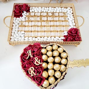 May include: A gold-colored rectangular tray with handles, filled with white candies and chocolate bars with gold lettering. The tray is decorated with burgundy roses. Below is a heart-shaped box with burgundy roses, gold-wrapped chocolates, and wooden letters.