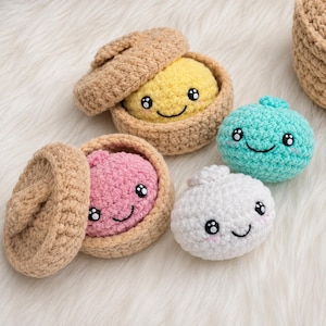 Puede incluir: Cuatro peluches de ganchillo con forma de albóndigas redondas y caras sonrientes. Dos están dentro de cestas de ganchillo color canela con tapa, mientras que una turquesa y una blanca están separadas, todas con ojos negros y mejillas rosadas.