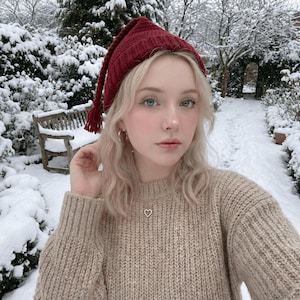 May include: A person wearing a burgundy knit hat with a tassel, a beige sweater, and a gold heart pendant necklace. The background shows a snowy outdoor scene with a wooden bench and snow-covered trees.