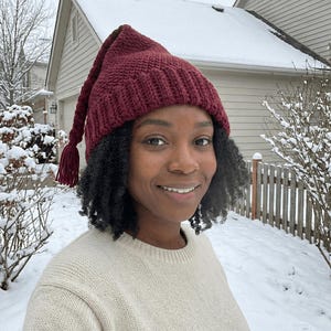 May include: A burgundy crochet hat with a long, braided tassel. The hat is worn by a person in a snowy outdoor setting. The hat has a pointed top and a textured knit pattern. The person is wearing a cream-coloured sweater.