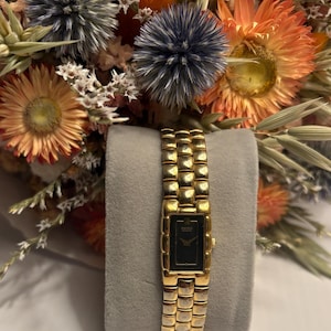 May include: A gold-toned wristwatch with a rectangular black face and a linked band. The watch is displayed on a gray surface, with a backdrop of dried flowers in shades of orange, blue, and white.
