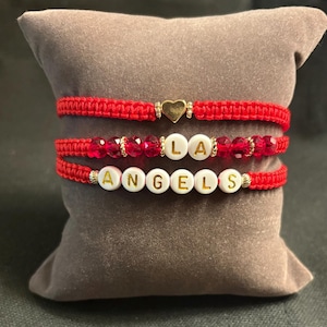 May include: Three red macrame bracelets. One features a gold heart charm, the second has red beads spelling "LA", and the third spells "ANGELS" with white letter beads. The bracelets are displayed on a gray pillow.