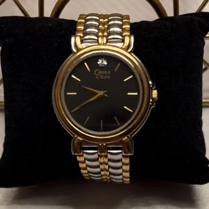 May include: A gold and silver-toned Caravelle by Bulova wristwatch with a black face and gold hands. The watch has a small diamond at the 12 o'clock position and is displayed on a black velvet pillow.