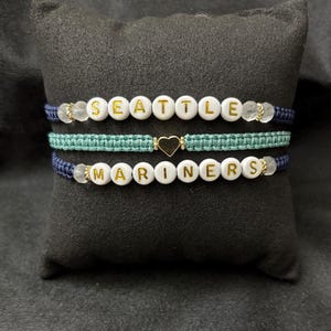 May include: Three handmade bracelets. One bracelet spells "SEATTLE" in gold letters on white beads, another is teal macrame with a gold heart, and the third spells "MARINERS" in gold letters on white beads. All are on a black pillow.